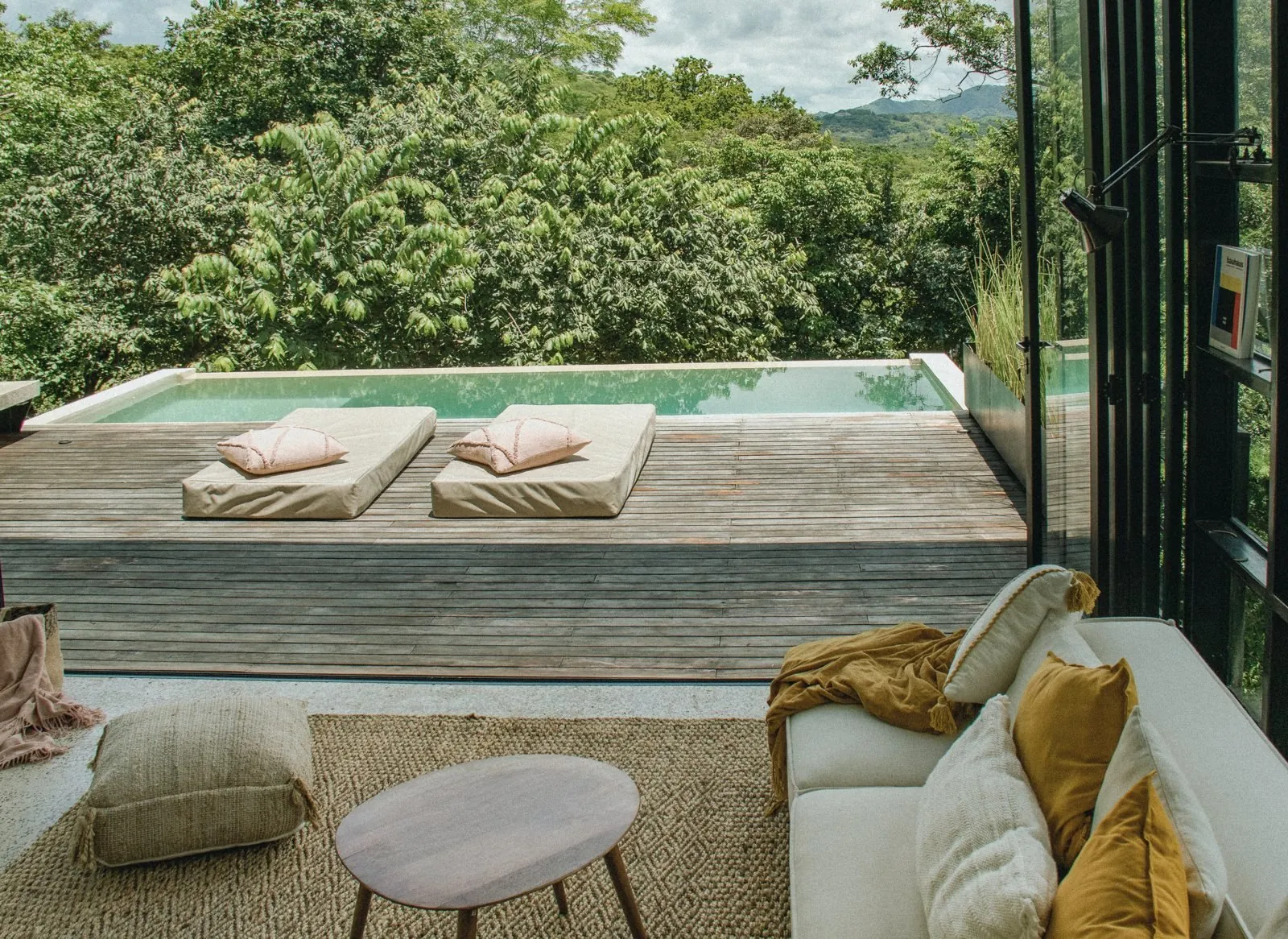 Desde el sofá: el deck abierto con dos cojines rosados, la piscina y las copas de los árboles, colinas a lo lejos.